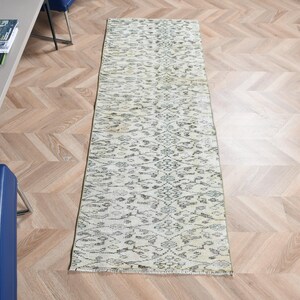 May include: A long, rectangular runner rug featuring a cream-colored base with an intricate, repeating floral and vine pattern in faded black and charcoal gray tones. The rug has a low-pile texture and finished edges along the short ends.