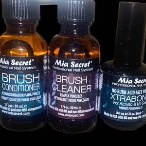Puede incluir: Tres botellas de productos para el cuidado de las uñas Mia Secret. Las botellas son negras con letras plateadas y contienen un líquido transparente. Las botellas están etiquetadas como "Brush Conditioner", "Brush Cleaner" y "No-Burn Acid-Free Primer Xtrabond for Acrylic & UV Gels".