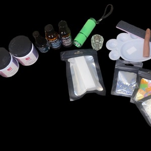 Könnte beinhalten: Ein Set an Nagelkunstzubehör, darunter eine Nagelfeile, ein Finger-Übungswerkzeug, eine kleine Glasschale, ein Nagellackentferner, eine Flasche Nagelkleber, eine Flasche Top Coat, eine Flasche Base Coat, eine Flasche Nagellack, eine Tüte mit weißen Nagelspitzen, eine Tüte mit Strasssteinen, eine Tüte mit bunten Nagel-Anhängern und eine Tüte mit weißen Perlen.