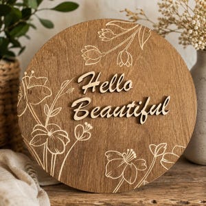 Puede incluir: Un letrero circular de madera con acabado marrón medio que presenta arte lineal floral grabado con láser. Las palabras Hello Beautiful se muestran en el centro en una fuente cursiva tridimensional en relieve de madera clara.