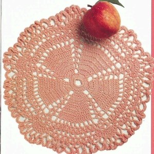May include: A delicate, crocheted doily in a soft pink color. The doily is circular and features a detailed, intricate pattern. A peach sits on top of the doily.