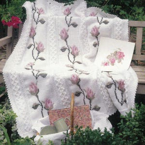 Pode incluir: Cobertor de crochê branco com um padrão de ponto alto e rosas cor-de-rosa bordadas. O cobertor está drapeado sobre um banco de madeira com uma renda de crochê branca e uma cesta com um livro e canecas.