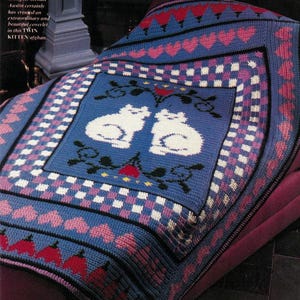 May include: A blue crocheted blanket with a white and pink checkered border. The center of the blanket features two white kittens with a red heart and floral design. The text "Designer Renate Austin certainly has created an extraordinary and beautiful coverlet in this TWIN KITTEN afghan." is visible in the top left corner of the image.