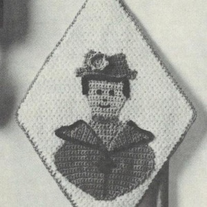 May include: A crocheted wall hanging in a diamond shape, featuring a detailed image of a person wearing a hat and coat. The design is in shades of white and dark gray, with a loop at the top for hanging.