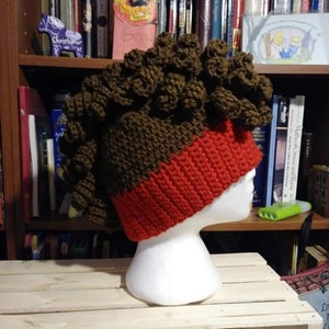 May include: A brown and red crocheted hat with a curly brown top. The hat is on a white mannequin head.
