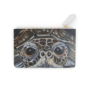 May include: A rectangular clutch featuring a detailed painting of a sea turtle's face. The turtle's eyes are large and dark, with a light-colored snout and a textured shell pattern. The clutch has a white zipper pull.