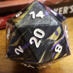 May include: A black, gold, and purple 20-sided die with white numbers. The die is made of a translucent material and has a glittery finish.