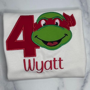 May include: White t-shirt with a red number 4 and a green cartoon turtle face with a red mask. The name "Wyatt" is embroidered in red script below the turtle. The shirt is likely for a child's birthday.