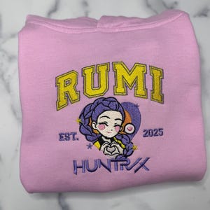 May include: A light pink hoodie with the word "RUMI" in yellow block letters. Below the word "RUMI" is a cartoon graphic of a girl with purple hair. The text "EST. 2025" and "HUNTRX" are also visible.