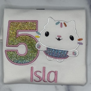 May include: A white t-shirt featuring an embroidered design of the number 5 in a glittery rainbow gradient with a pink outline, alongside a white cat character with colorful sprinkles and a blue glittery skirt, with the name Isla in pink text.