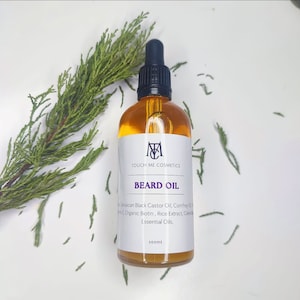 May include: A clear glass bottle with a black dropper top and a white label. The label says "TOUCH ME COSMETICS" and "BEARD OIL". The bottle contains a yellow liquid. The ingredients listed on the label are Jamaican Black Castor Oil, Comfrey Oil, Organic Biotin, Rice Extract, Calendula, and Essential Oils. The bottle is 100 ml.