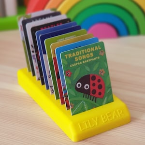 May include: A yellow card holder with the words "Lily Bear" printed on the front. The holder is filled with colourful cards featuring different song titles, including "Traditional Songs" by Caspar Babypants.