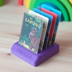 May include: Purple plastic book holder with the name Amelia on the front. The holder is holding a stack of children's books, including "The Gruffalo" and "My First"