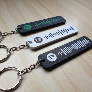 May include: Three rectangular keyrings featuring a Spotify code design. One is black with a green Spotify logo, another is white with a black Spotify code, and the third is black with a white Spotify logo. Each keyring has a silver chain and ring.
