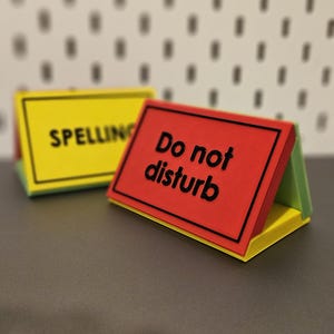 May include: Two triangular desk signs with raised black lettering. The front sign is red with the text "Do not disturb" in black. The sign behind it is yellow with the text "SPELLING" in black. Both signs feature a black border frame.