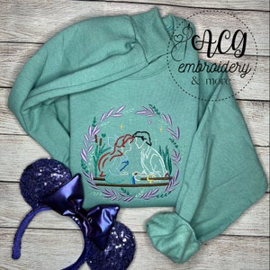May include: A teal green sweatshirt with an embroidered design of a couple kissing. The design is surrounded by a wreath of leaves and flowers. The couple is depicted in a romantic setting with stars in the background.
