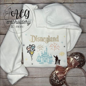 May include: White sweatshirt with embroidered Disneyland text and a colorful image of Sleeping Beauty's castle, fireworks, Mickey Mouse balloons, and a silhouette of a person holding a child's hand.
