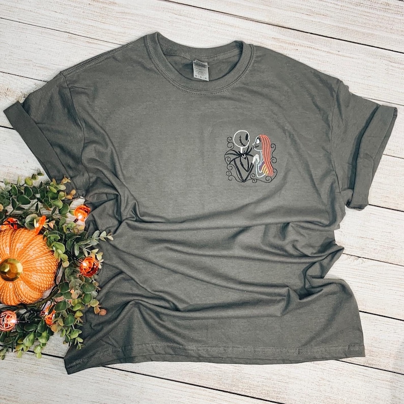 Puede incluir: Una camiseta gris oscuro con una imagen bordada de Jack Skellington y Sally de Pesadilla antes de Navidad.