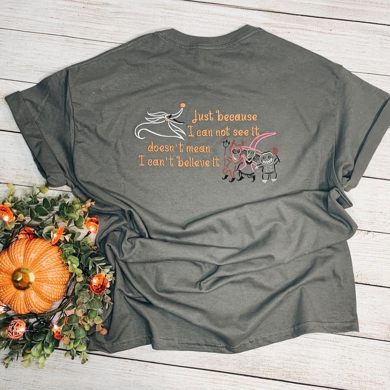 Puede incluir: Una camiseta verde oscuro con un dise&ntilde;o bordado con personajes de la pel&iacute;cula "Pesadilla antes de Navidad". El texto dice "Solo porque no pueda verlo no significa que no pueda creerlo."