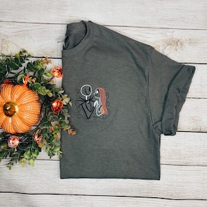 Puede incluir: Una camiseta gris oscuro con una imagen bordada de Jack Skellington y Sally de Pesadilla antes de Navidad.