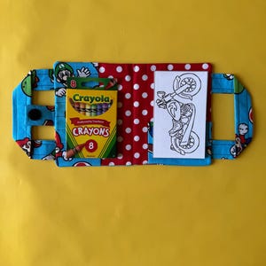 May include: A blue fabric case with a cartoon character print, holding a box of Crayola crayons and a black and white motorcycle coloring page. The case has a red polka dot interior and a button closure.