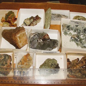 Puede incluir: Una colección de diversos especímenes minerales en una caja de madera. Los minerales están etiquetados con sus nombres, incluyendo Calcita, Byssolite Tremolite y Talco verde. Los especímenes están dispuestos en pequeñas cajas blancas con una regla para la escala.