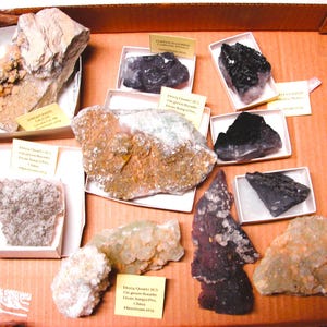 May include: A collection of various mineral specimens in a cardboard box. The specimens are labeled with their names and locations. The minerals include fluorite, quartz, and calcite.