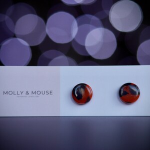 May include: A pair of small, round stud earrings with a black and orange marbled design. The earrings are displayed on a white card with the text "MOLLY & MOUSE HANDMADE JEWELLERY".
