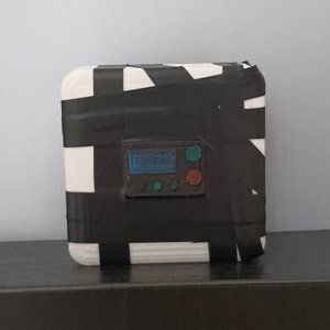 Puede incluir: Una caja blanca envuelta con cinta adhesiva negra. La caja tiene una pequeña pantalla digital y un botón.