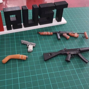 Könnte beinhalten: Ein 3D-gedrucktes Schild mit der Aufschrift "RUST" und einem roten und weißen Symbol daneben. Auf einer grünen Oberfläche liegen vier Spielzeugpistolen.