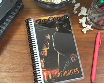 May include: A spiral bound notebook with a clear plastic cover featuring the movie poster for the Western film "Unforgiven". The poster features Clint Eastwood, Gene Hackman, Morgan Freeman, and Richard Harris. The notebook is on a wooden surface with a VHS tape and a bowl of popcorn.