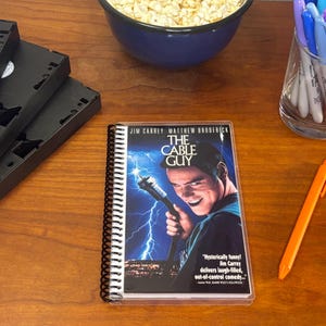Puede incluir: Una libreta de espiral con una cubierta que presenta el cartel de la película "The Cable Guy" protagonizada por Jim Carrey y Matthew Broderick. El cartel presenta una imagen en blanco y negro de Jim Carrey sosteniendo un cable con un rayo en el fondo. El texto "Hysterically funny! Jim Carrey delivers laugh-filled, out-of-control comedy..." está impreso en la parte inferior del cartel.