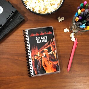 Op de afbeelding: Een spiraalgebonden notitieboek met een doorzichtige plastic hoes met de DVD-hoes van de film "Ocean's Eleven" met George Clooney, Brad Pitt, Matt Damon en Andy Garcia. De DVD-hoes is zwart met witte tekst en toont de acteurs in een scène uit de film.