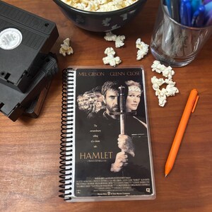 Puede incluir: Una libreta de espiral con una portada de póster de película que presenta a Mel Gibson y Glenn Close en la película Hamlet. El póster tiene un fondo azul oscuro con el título Hamlet en texto blanco. Los actores sostienen espadas y visten ropa de época.