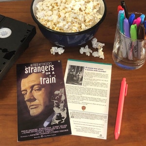 Puede incluir: Una cinta VHS, un tazón de palomitas de maíz, un vaso de marcadores, un bolígrafo rosa y un cartel de película para la película de Alfred Hitchcock "Extraños en un tren". El cartel presenta una ilustración en blanco y negro de Farley Granger, el personaje principal de la película. El texto del cartel dice "Alfred Hitchcock's Strangers on a Train".
