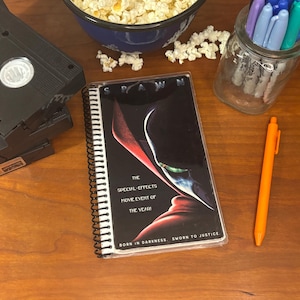 May include: A black and red spiral bound notebook with a clear plastic cover featuring a movie poster for the film "Spawn". The poster features a red and black character with green eyes. The text on the poster reads "The Special-Effects Movie Event of the Year! Born in Darkness. Sworn to Justice."
