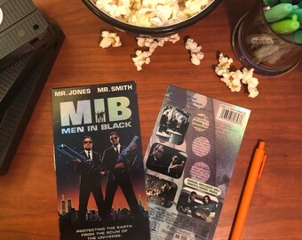 May include: A VHS tape case for the movie Men in Black featuring Will Smith and Tommy Lee Jones. The case is black and has a silver holographic back with the movie title and a description of the movie. The case is sitting on a wooden table with a bowl of popcorn and a pen.