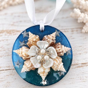 May include: A circular blue resin ornament featuring a central white flower with pearl accents, surrounded by small brown spiral seashells and clear star-shaped embellishments. The ornament hangs from a white satin ribbon loop.