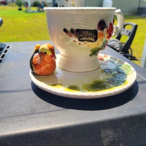 Può includere: Un set di tazza e piattino in ceramica color crema con una statuetta tridimensionale di anatra mandarina sul piattino, un motivo dipinto di anatra e fiori sulla tazza e un dettaglio del bordo a corda sia sulla tazza che sul piattino.
