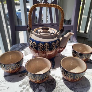 Puede incluir: Un juego de té de cerámica con una tetera y cuatro tazas a juego. La tetera tiene un asa trenzada y está decorada con diseños de hojas azules, rojas y doradas. Las tazas tienen un diseño similar. El juego está sobre un mantel con estampado floral.