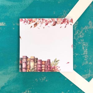 May include: A square notepad featuring a watercolour illustration of a stack of books and autumnal leaves. The notepad is white with a decorative border of books and leaves. The background is teal with a white frame and a piece of paper.