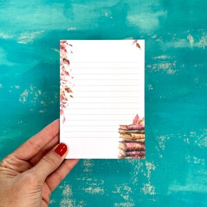 May include: A white lined notepad with a watercolor design of autumn leaves and a stack of books. The notepad is held by a hand with red nail polish. The design features shades of red, orange, and brown.