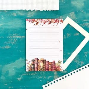 Könnte beinhalten: Ein liniertes Notizbuch mit einer Aquarellillustration eines Bücherstapels und Herbstblättern. Die Oberseite des Notizbuchs zeigt einen Rand aus roten und orangen Blättern. Das Notizbuch liegt auf einer türkisfarbenen Oberfläche mit anderen Papierelementen.