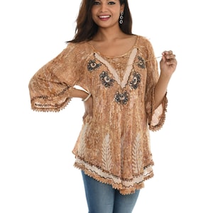 May include: A tan-colored tunic top with a floral design around the neckline and sleeves. The top has a loose, flowing fit with bell sleeves and a decorative trim along the hemline. The fabric has a textured appearance, and the top is paired with blue jeans.