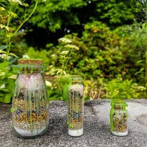 May include: Three glass bottles with cork stoppers filled with dried herbs and flowers. The bottles are decorated with green wax drippings.