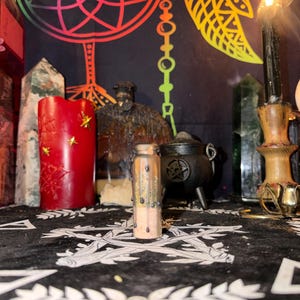 May include: A spiritual altar setup featuring a red candle with gold star accents, a small glass bottle with pink contents, and a black cauldron. A burning black candle and crystals are also present.