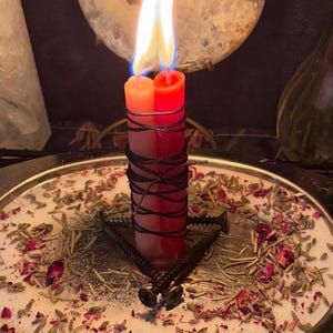 Pode incluir: Duas velas unidas, uma laranja e uma vermelha, estão acesas com chamas visíveis. As velas estão envolvidas por um fio preto fino e rodeadas por três parafusos de metal preto dispostos em triângulo num prato prateado com ervas e pétalas.