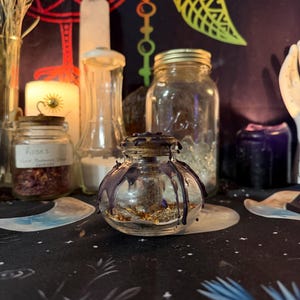 May include: A small, clear glass jar with a dark purple wax seal and drips. The jar contains dried herbs and is surrounded by other jars, candles, and decorative items on a patterned surface. The scene evokes a mystical or spiritual theme.