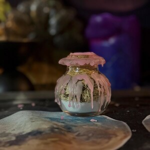 May include: A small, clear glass jar with a cork stopper, featuring pink wax dripping down the sides and over the lid. The jar is filled with a layer of white powder at the bottom and a layer of dried green herbs or plant matter on top.