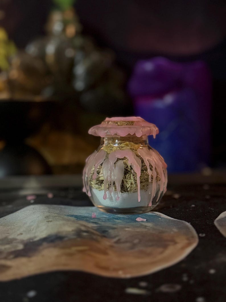May include: A small, clear glass jar with a cork stopper, featuring pink wax dripping down the sides and over the lid. The jar is filled with a layer of white powder at the bottom and a layer of dried green herbs or plant matter on top.